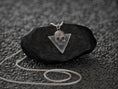 Load image into Gallery viewer, May include: A silver skull pendant necklace with a triangular frame. The pendant is hanging from a silver chain on a black rock.