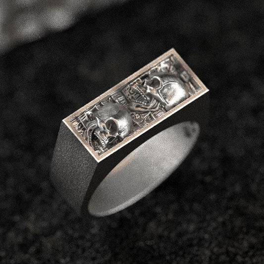 Three-quarter top-down view of the sterling silver bar signet ring showcasing two skull reliefs side by side on a rectangular face.