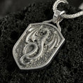 Load image into Gallery viewer, Polished silver coiled dragon shield necklace displayed on volcanic stone background with sharp sculpted relief