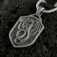 Load image into Gallery viewer, Oxidized coiled dragon shield pendant necklace photographed on volcanic rock with deeply engraved fantasy dragon detail