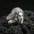 Load image into Gallery viewer, Polished silver Venus de Milo ring featuring serene goddess facial expression and flowing hair details, centered close-up on dark porous volcanic stone