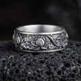 Load image into Gallery viewer, Oxidized lotus flower engraved band ring photographed on volcanic rock with detailed floral relief and artisan botanical texture