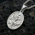 Load image into Gallery viewer, Polished silver Hela necklace pendant on volcanic rock, crisp engraved scene and textured background, close-up lighting