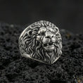 Load image into Gallery viewer, Oxidized sterling silver lion head ring on textured volcanic rock, macro close-up showing roaring expression, detailed mane curls, and sculpted fangs