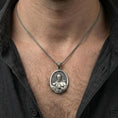 Load image into Gallery viewer, Oxidized sterling silver Saint Francis pendant worn on neck on a braided chain, centered chest framing, moody lifestyle studio shot