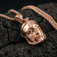 Load image into Gallery viewer, Rose gold plated Spartan helmet pendant on textured volcanic rock, luxury close-up capturing the raised faceplate design and warm metallic glow