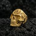 Load image into Gallery viewer, Gold plated skull ring with Masonic square and compass “G” emblem and ornate side engraving, macro close-up on textured volcanic rock