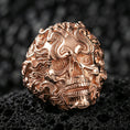 Load image into Gallery viewer, Rose gold plated ornate smoke skull ring with spiral eyes and wave-like scrollwork, close-up on volcanic rock