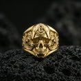 Load image into Gallery viewer, Gold plated Masonic skull ring with compass-and-G symbol and detailed baroque scroll patterns, close-up on volcanic rock background