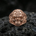 Load image into Gallery viewer, Sterling silver masonic skull ring with a rose gold plated finish, sculpted skull signet ring featuring triangle eye emblem and ornamental engraving, close-up on volcanic rock