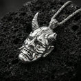 Load image into Gallery viewer, Sterling silver oni mask pendant in a polished silver finish, horned Japanese demon face with deep carved details, macro on volcanic stone