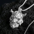 Load image into Gallery viewer, Polished silver oni mask pendant necklace on textured volcanic rock, crisp macro product photo emphasizing clean edges, sharp facial lines, and bright reflective highlights on the sculpted details.