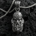 Load image into Gallery viewer, Oxidized Viking warrior head pendant necklace on textured volcanic rock, centered close-up highlighting the stern face, flowing beard, helmet rune detail, and deep antique shadowed contrast.