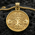 Load image into Gallery viewer, Gold plated Yggdrasil tree of life pendant necklace on textured volcanic rock, premium close-up with warm reflective shine across the tree roots, wolves, and intricate Nordic patterning.