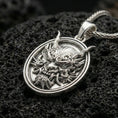 Load image into Gallery viewer, Polished silver demon mask medallion pendant necklace on textured volcanic rock, crisp macro product photo emphasizing clean edges, sharp relief lines, and bright reflective highlights on the sculpted details.