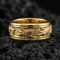 Load image into Gallery viewer, Gold plated wolf head band ring on textured volcanic rock, premium close-up with warm reflective shine across the wolf profiles, engraved fur lines, and raised ornamental contours.