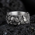 Load image into Gallery viewer, Oxidized skull and floral band ring on textured volcanic rock, centered close-up highlighting the raised skull accents, blooming flower relief, and deep antique shadowed contrast.