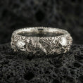 Load image into Gallery viewer, Polished silver skull and koi floral band ring on textured volcanic rock, crisp macro product photo emphasizing clean edges, sharp koi-and-flower engraving, and bright reflective highlights across the sculpted band.
