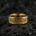 Load image into Gallery viewer, Gold plated geometric knotwork band ring on textured volcanic rock, premium close-up with warm reflective shine across the repeating interlace motif and raised contours.