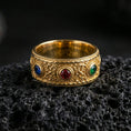 Load image into Gallery viewer, Gold plated ornate gemstone band ring on textured volcanic rock, premium close-up showcasing warm gold tone over the floral scroll pattern and the three colorful stones in sharp focus.