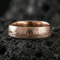 Load image into Gallery viewer, Rose gold plated Damascus-style band ring on volcanic stone, refined macro shot with soft rosy highlights and pronounced wavy layered texture.