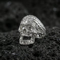 Load image into Gallery viewer, Polished silver ornate skull ring with carved scrollwork filigree and rope-edge band on porous volcanic stone, close-up highlighting crisp engraving and shine