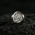 Load image into Gallery viewer, Polished silver raven signet ring featuring engraved crest details and rounded face, close-up studio shot on porous volcanic stone