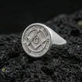 Load image into Gallery viewer, Polished silver Freemason signet ring featuring square and compass engraving with circular motto border, close-up on porous volcanic stone