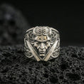Load image into Gallery viewer, Polished silver horned demon face ring featuring twin daggers and engraved side panels, close-up product shot on volcanic stone