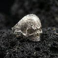 Load image into Gallery viewer, Polished silver gothic skull signet ring featuring ornate floral filigree carving across the band, close-up on volcanic stone
