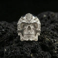 Load image into Gallery viewer, Polished silver cyber skull ring featuring mechanical detailing, hollow eye sockets, and sharp contour lines, close-up on volcanic stone
