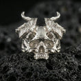 Load image into Gallery viewer, Polished silver horned skull statement ring with textured curved horns and sculpted skull face, close-up on volcanic stone