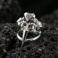 Load image into Gallery viewer, Polished silver heart dagger ring featuring a sculpted blade and three heart motifs, close-up product shot on volcanic stone