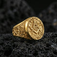 Load image into Gallery viewer, Gold plated Masonic signet ring featuring square and compass “G” emblem with engraved floral scroll band, close-up on volcanic rock