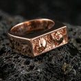 Load image into Gallery viewer, Sterling silver minimalist signet ring with a rose gold plated finish, rectangular cutout face with skull-inspired relief accents, close-up on volcanic rock