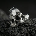 Load image into Gallery viewer, Sterling silver shattered skull ring in a polished silver finish, sculpted skull face with broken-style edges and bold unisex profile, close-up on volcanic stone