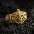 Load image into Gallery viewer, Sterling silver Masonic skull ring with a gold plated finish, Freemason-inspired skull design with eye-in-triangle motif and rich carved textures, macro on volcanic rock