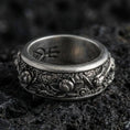 Load image into Gallery viewer, Oxidized sterling silver lotus spinner band ring on dark volcanic rock, raised lotus flowers and scrollwork on rotating center, premium macro product shot.