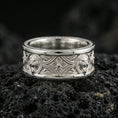 Load image into Gallery viewer, Polished silver lion crest band ring on black volcanic stone, crisp lion head relief medallions with geometric cross motif and ornate lines, centered close-up studio lighting.