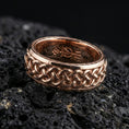 Load image into Gallery viewer, Rose gold plated braided Celtic knot band on black volcanic stone, intricate interlace pattern with warm reflective highlights, refined product close-up.