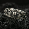 Load image into Gallery viewer, Oxidized sterling silver gothic skull gear band ring on black volcanic stone, darkened relief highlighting the central skull, side scrollwork, and industrial gear details, macro product photo.