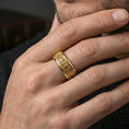 Load image into Gallery viewer, Gold plated fleur de lis ring worn on a finger, warm reflective finish emphasizing the raised pattern and bold band profile.