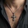 Load image into Gallery viewer, Oxidized Chicano cross pendant with rose motifs worn on the neck in a lifestyle shot, styled against an open dark shirt for a bold statement look
