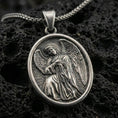 Load image into Gallery viewer, Oxidized angel pendant with oval frame and detailed winged figure displayed on dark volcanic rock in a close-up luxury jewelry composition