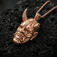 Load image into Gallery viewer, Sterling silver oni mask pendant with a rose gold plated finish, horned Japanese oni face with fangs and sculpted contours, macro on black volcanic rock