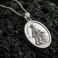 Load image into Gallery viewer, Polished silver Saint Rochus pendant with oval frame, devotional figure, and dog relief placed on textured volcanic rock in a premium studio product shot