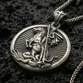 Load image into Gallery viewer, Oxidized Saint George pendant with mounted warrior relief, spear detail, and round medallion design displayed on dark volcanic rock in a close-up luxury jewelry composition