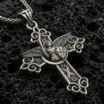 Load image into Gallery viewer, Oxidized Saint Michael cross pendant with winged archangel figure, shield detail, and ornate gothic scrollwork displayed on dark volcanic rock in a close-up luxury jewelry composition