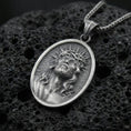 Load image into Gallery viewer, Oxidized Jesus Christ pendant with crown of thorns, radiant halo detail, and sacred portrait displayed on dark volcanic rock in a close-up luxury jewelry composition