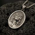 Load image into Gallery viewer, Bronze Jesus Christ pendant with crown of thorns, radiant halo detail, and sacred portrait displayed on dark volcanic rock in a close-up luxury jewelry composition